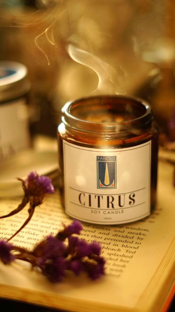 A citrus-scented soy candle rests atop an open book alongside some purple flowers