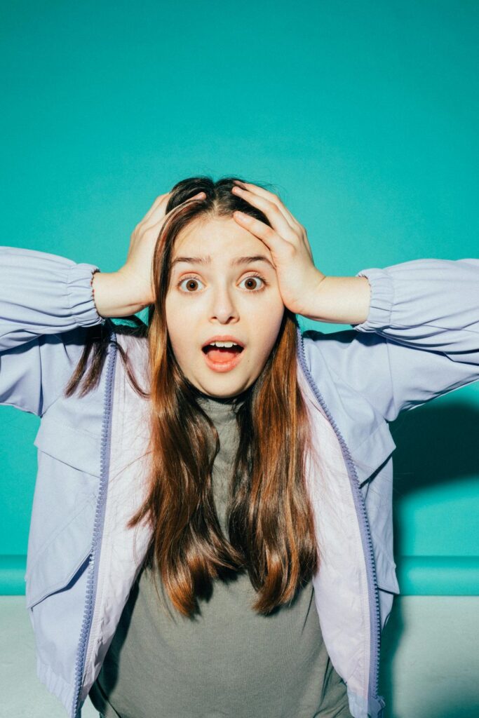 A teenage girl has a surprised look on her face and holds her hands against the sides of her head in a gesture of overwhelm