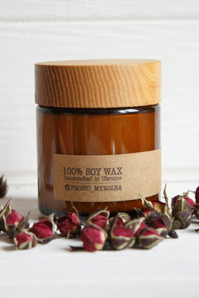 A candle in a brown glass jar with a label that reads '100% soy wax handcrafted in Ukraine' with scattered dried rose buds in front of it