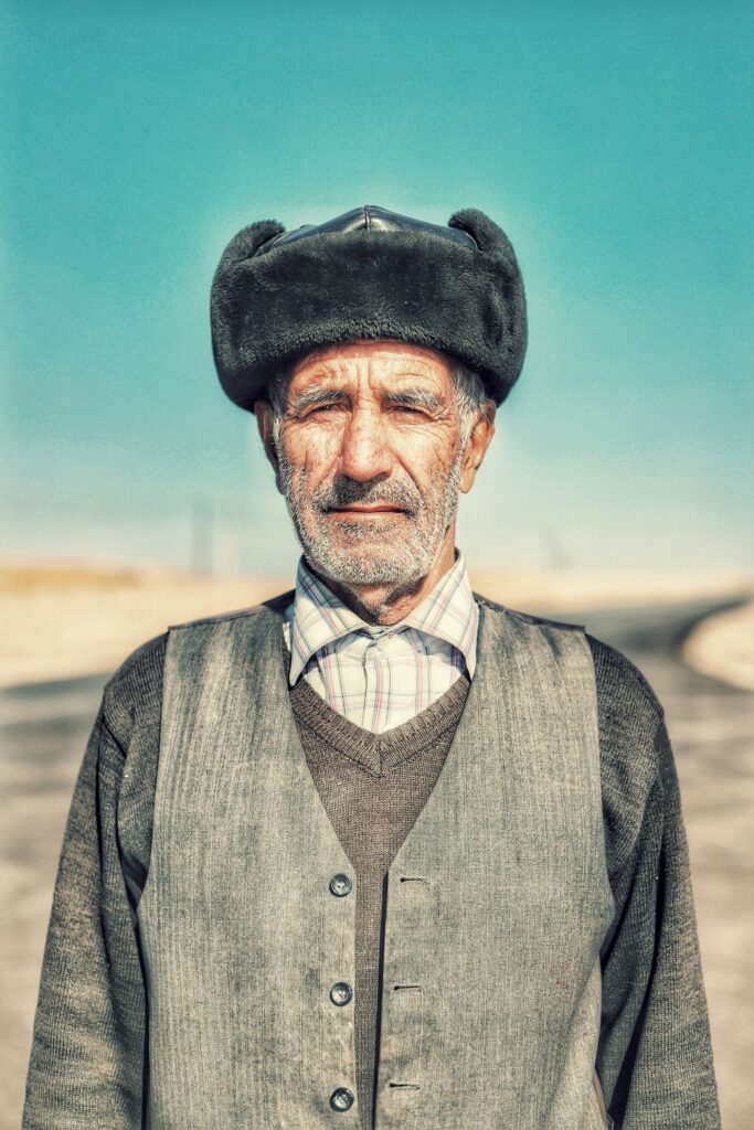 An older man wears a light plaid collared shirt buttoned at the very top under a gray V-neck sweater and an open gray button-up vest, with a gray ushanka (Russian hat)