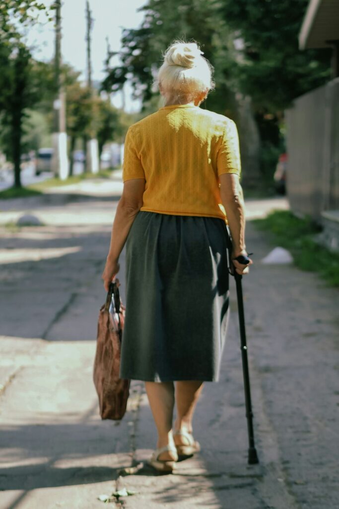 An older woman seen from behind walking up a sidewalk with a cane, carrying a large brown handbag, and wearing a calf-length dark coloured skirt with a buttercup yellow short-sleeved sweater