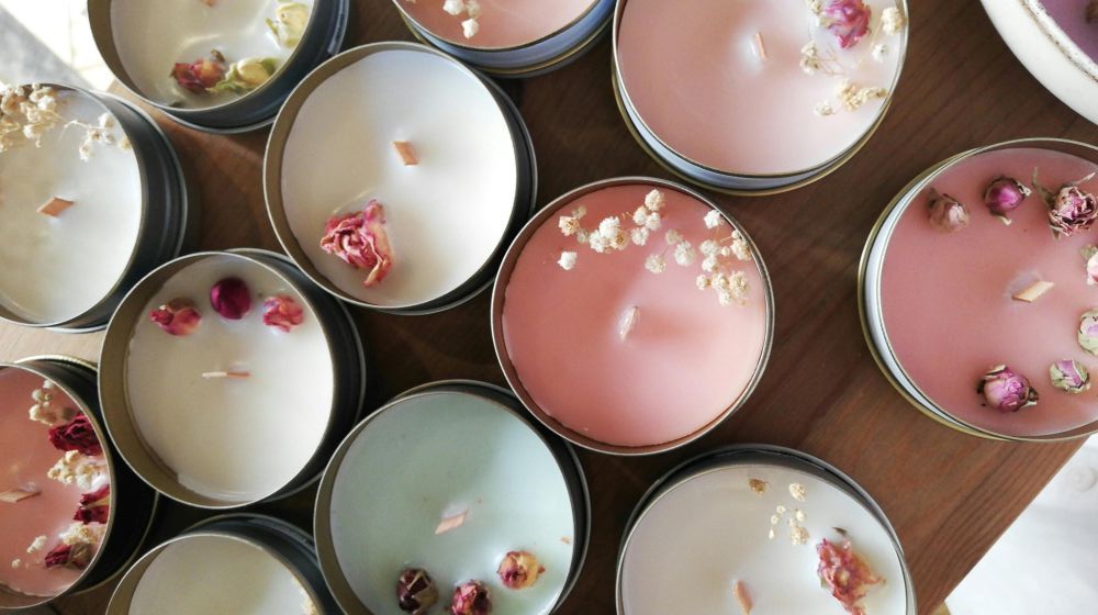 Top-down close-up photo of several pastel-coloured clean burning candles in tin jars with woodwicks and dried flowers