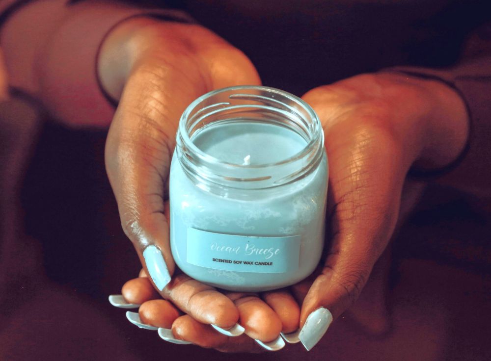 Close-up photo of a woman's hands with light blue-painted fingernails holding a small candle with matching light blue wax in a clear glass jar with a label that reads 'Ocean Breeze scented soy wax candle' (clean burning candles made in Kampala, Central Region, Uganda)
