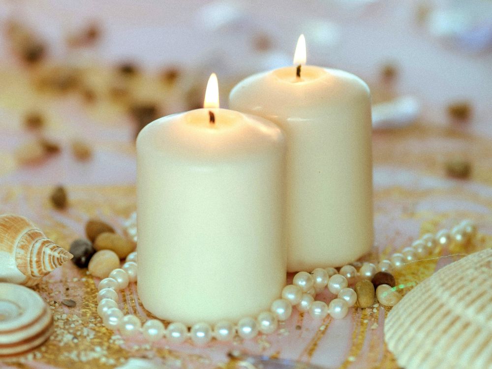 Close-up photo of an oceanic vignette featuring two off-white coloured lit pillar candles amidst a strand of white pearls, various seashells, and gold glitter