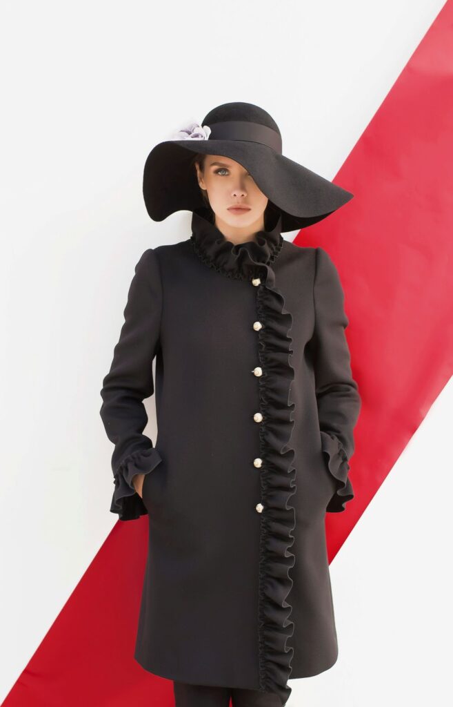 A woman poses in front of a white wall with a large red diagonal stripe wearing a smart long black structured jacket with white buttons, a ruffled collar and vertical hemline down opening and ruffled sleeves, and a large floppy black hat