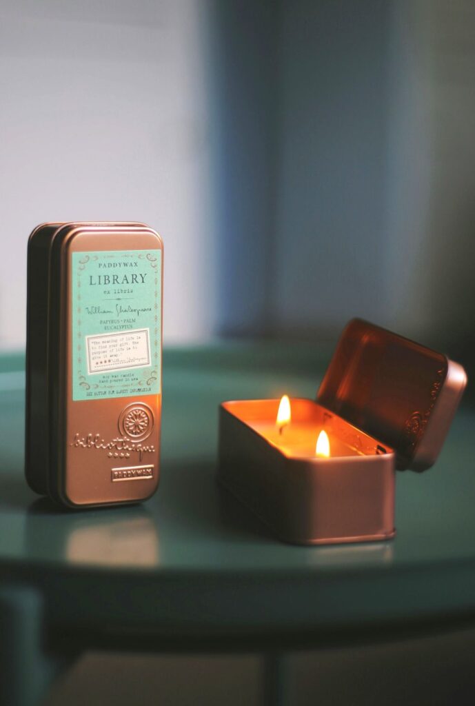 Two clean burning candles (of the same kind) poured in rectangular-shaped rose brass-coloured tin containers rest on a table. One stands upright with the lid on (showing the label, which reads 'library' and 'soy wax candle hand poured in usa') and the other lays flat with the lid open and the two wicks lit, producing a pleasant glow that reflects off the metal tin container
