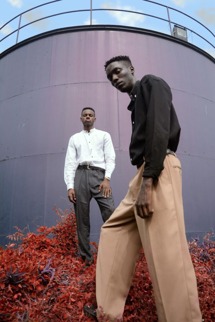 Two smartly-dressed men pose in front of a large structure and blue sky. The man closest to the camera wears tan slacks with a black button-down shirt tucked in, and the man further back wears gray slacks with a belt and a white button-down shirt tucked in