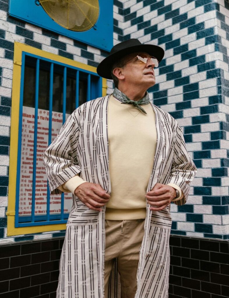 Fashion is important no matter one's age, sex, or gender identity: an older man wearing tinted glasses, a black fedora, a muted teal bandana tied around his neck, a cream-coloured sweater, khakis, and a long open jacket with a white and black striped print poses in front of a colourful building with alternating white and black bricks and bright blue painted bars in front of a yellow-framed window