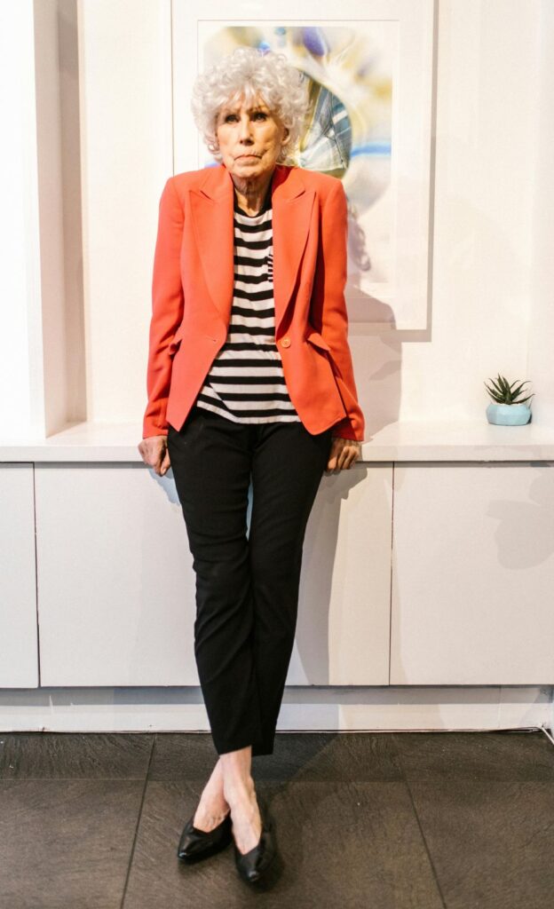 A smartly-dressed older woman leans against a ledge in front of framed wall art, wearing an open orange blazer (perfect colour pop) over a loosely fit black and white-striped shirt, fitted black pants, and black flats