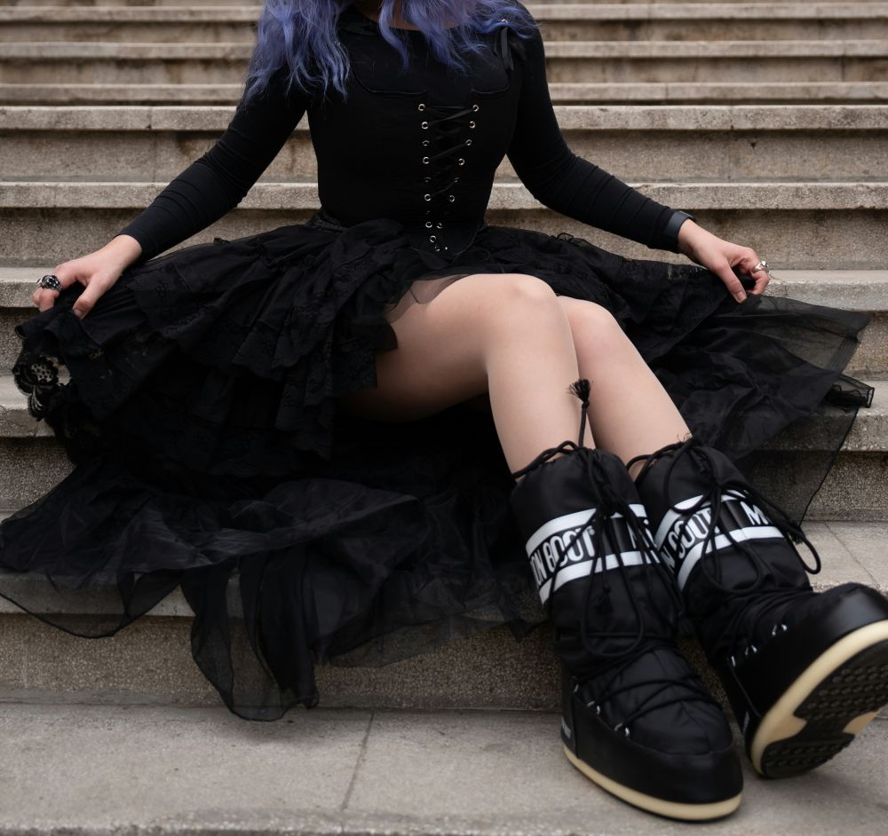 Close-up photo of a woman seated on stone steps in a black dress with a lace-up boddice-style long-sleeved top and large lacy/tulle-style skirt with funky chunky poofy black platform snow boots, inspiring Babymetal-style classy Halloween costumes