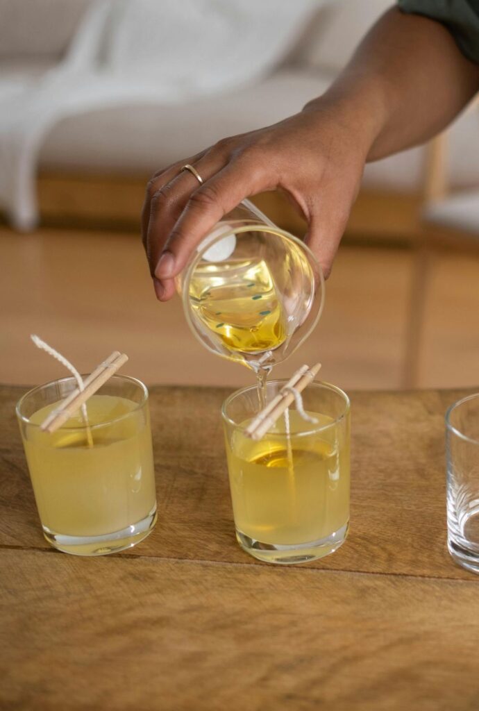 Close-up photo of a person making clean burning candles by pouring a clear/gold-coloured wax from a glass container into votive-sized clear glass candleholders with wicks secured down the center, hanging from small sticks spanning the top rim