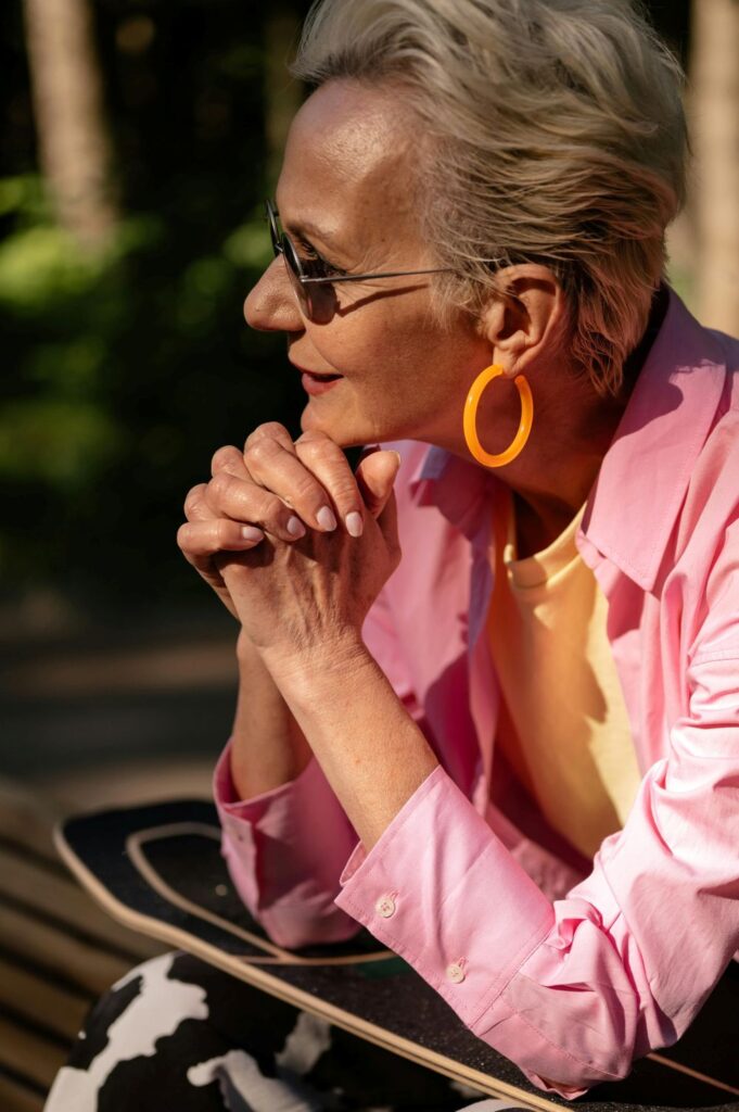 An older woman is photographed wearing an open light bubblegum pink blouse over a yellow shirt0 and large bright orange hoop earrings, with black and white cow-print pants. A refreshing reminder that fashion is important for everyone no matter their age