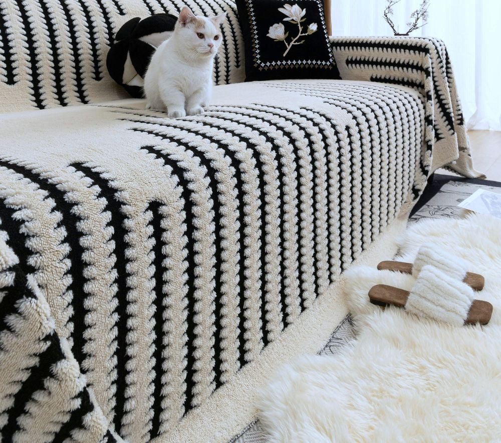 A textured B&W striped slipcover is draped over a sofa in front of a white shag rug with a white kitten on top of the sofa, and black throw pillows