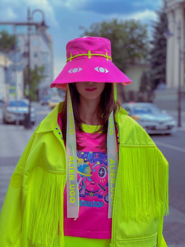 A woman's outfit happily screams that fashion is important to her: she wears a baggy open bright neon yellow fringe jacket over a neon pink shirt with a colourful illustration on the front and neon yellow neckline and undershirt. Her eyes are covered by the large low-hanging brim of her floppy neon pink hat with white eyes on the front (approximately where her own eyes are, unseen underneath). A neon yellow cord wraps around the circumference of the hat. A brilliant ensemble