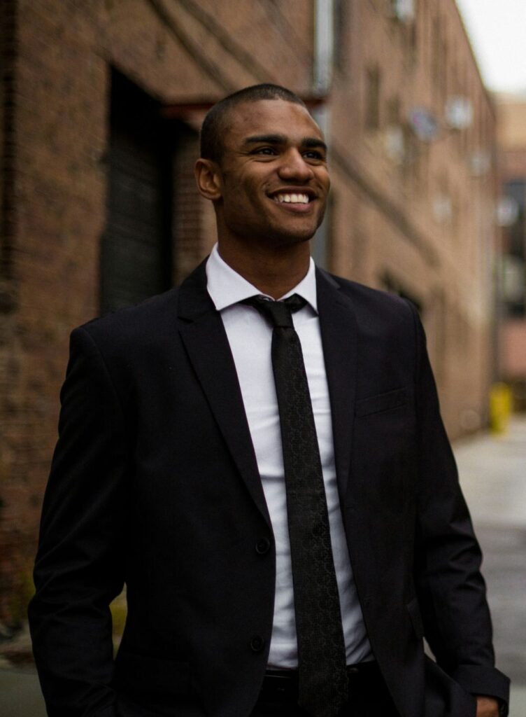 A man wearing a black suit jacket, white collared shirt underneath and black tie smiles while walking between buildings