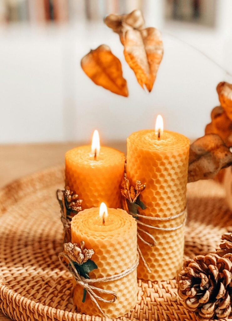 Winter-themed vignette of three lit beeswax pillar candles with honeycomb-indented edges and tied with twine and small gold-painted sparkly mistletoe sprigs on a round wicker tray next to a pinecone. Beeswax candles are some of the best clean burning candles