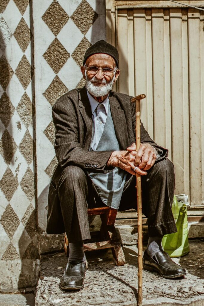 Fashion is important to people of all ages, in all places: an older gentleman sits with his cane in his hands on a small low stool in front of a building with a white diamond print on the wall, wearing a brown pin-striped suit, brown cap, and denim blue button-down shirt underneath, with black dress shoes and socks