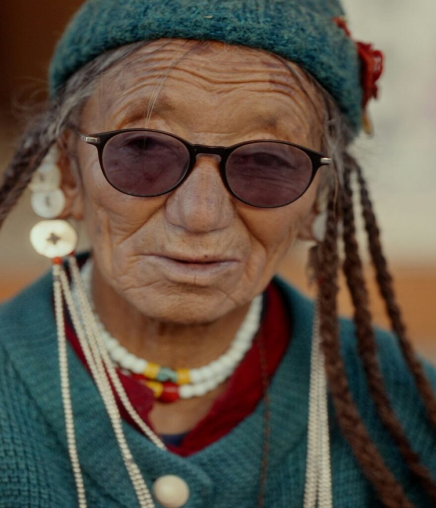 Fashion is important to all of us, including the older woman in this close-up photograph wearing a teal knit button-up sweater and matching knit teal hat, with long dreadlocks, tinted glasses, large earrings, and a white beaded necklace