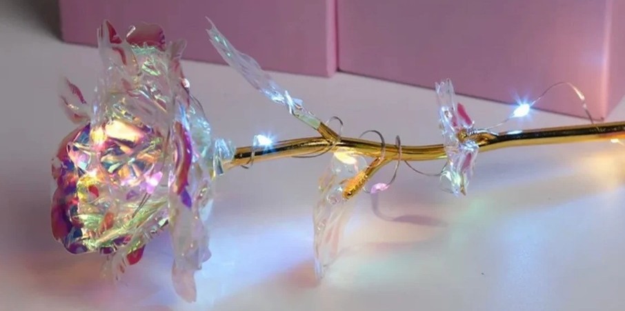 An iridescent artificial rose with gold stem lies on its side with a strand of fairy lights wound around it for extra sparkle