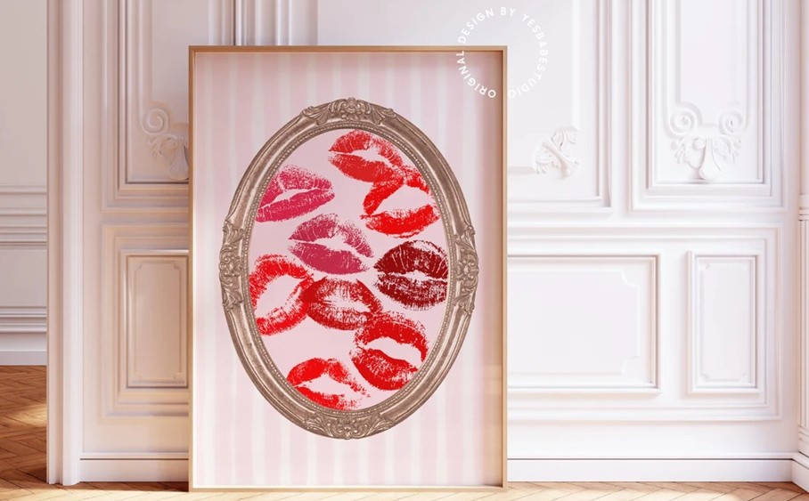 A framed art print (featuring a pale pink and white striped background and gilded oval-shaped frame around several pink and red lipstick kisses) rests against a wall.