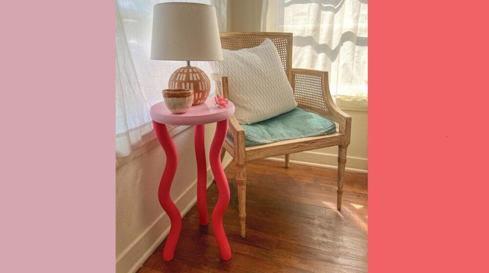 A small table with three squiggly dark coral-coloured legs and pale pink round top stands in the corner of a sunny room next to a wicker chair