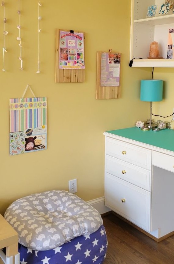 In the corner of the study is one end of a built-in desk below built-in shelving. A blue stick lamp is in the corner amidst pretty decorations, and a plush animal bag stuffed with blankets with a cushion on top is on the floor next to it, beside a coffee table for toddler artwork. Colourful collages hang on the wall above, filling the corner with beauty and calm