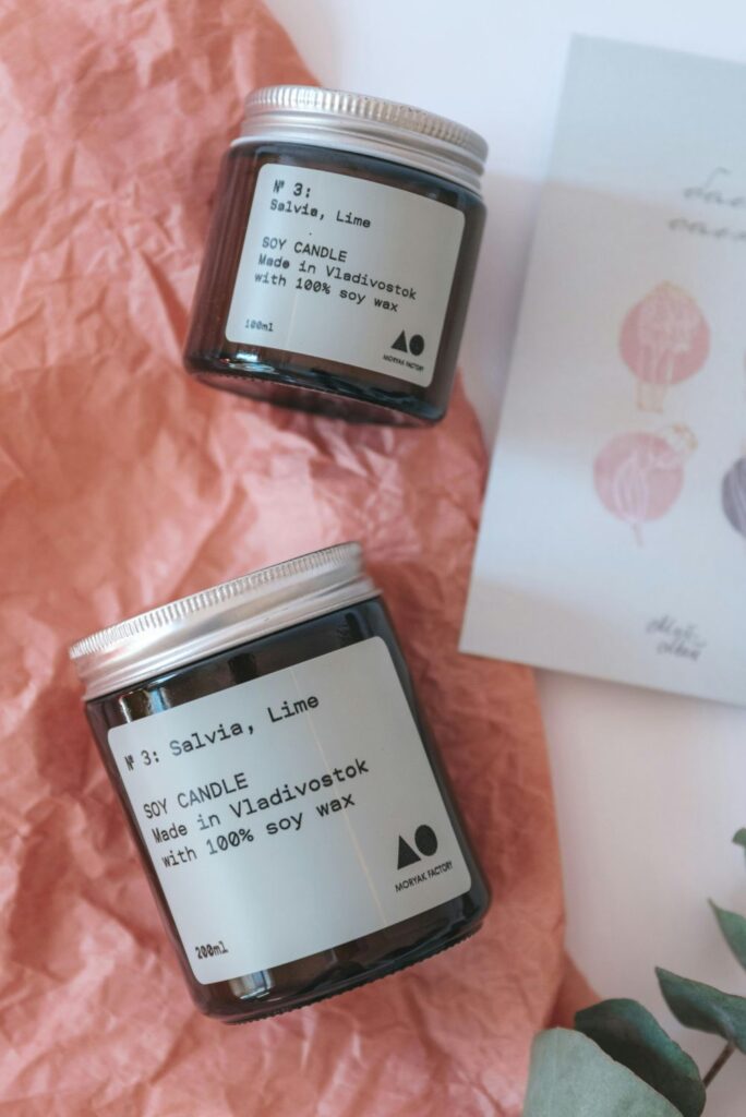 Two clean burning candles in brown glass jars lie on their side atop peach tissue paper next to a card, with labels that read 'Salvia, Lime' and 'soy candle made in Vladivostok with 100% soy wax'