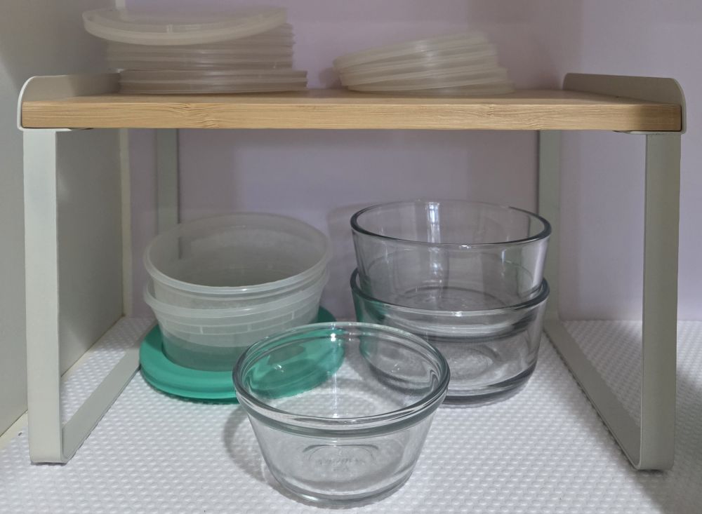 Kitchen household products are some of the most useful! The bamboo cabinet shelf organizer seen close-up here allows small glass food storage containers to stack underneath alongside plastic containers and lids, with piles of lids on top.