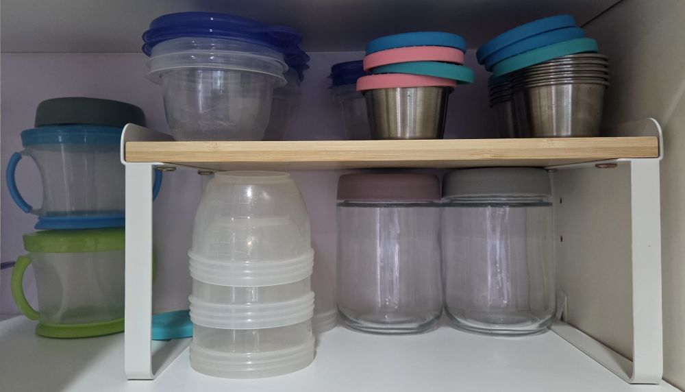 Kitchen household products like the attractive and simple cabinet shelf organizer depicted here help organize what would otherwise be an overcrowded kitchen. Glass jars with lids as well as a stack of plastic food storage cups fit perfectly underneath while small metal cups and lids and other plastic containers and lids fit stacked on top.