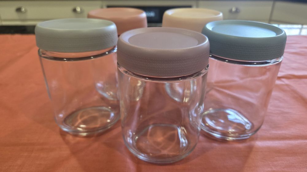 Five glass food/beverage storage jars atop a counter with different coloured lids in soft muted 'coastal colours'