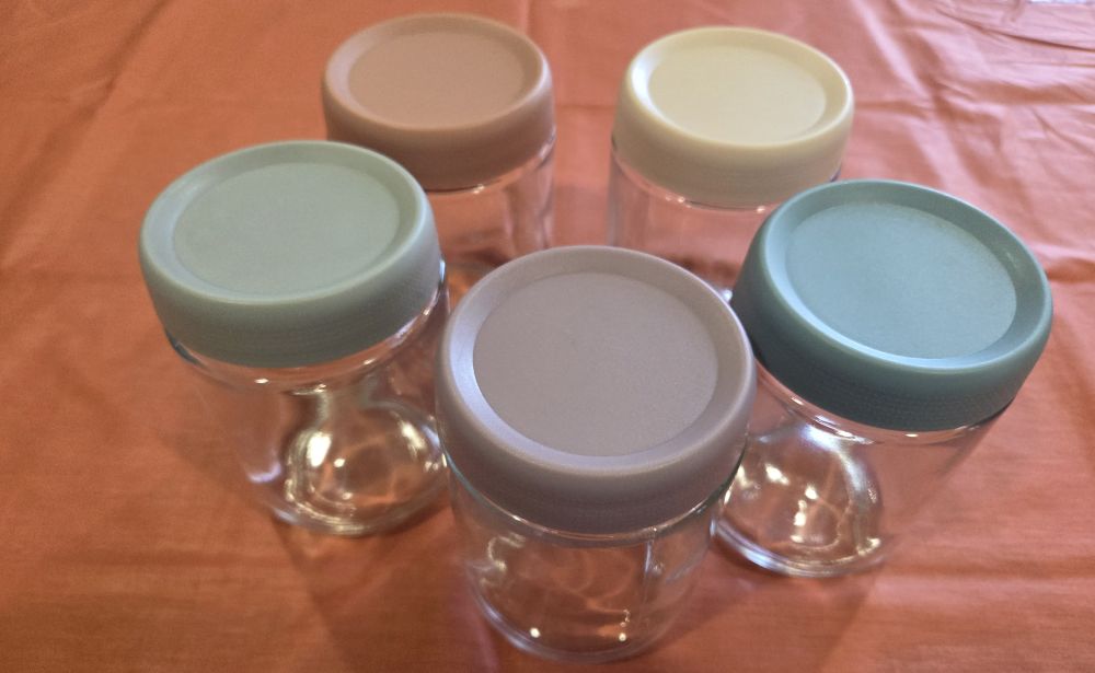 Five glass food/beverage storage jars atop a counter with different coloured lids in soft muted 'coastal colours' seen from top down