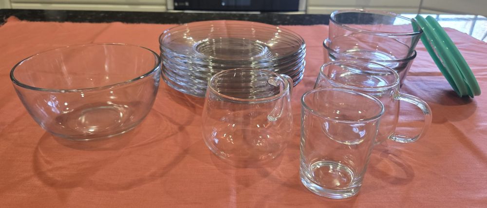 Many different clear glass dishes (including plates, food storage containers, etc.) sit atop coral coloured fabric on a kitchen counter. It's important to use non-toxic household products like these in the kitchen to avoid eating hot foods from plastic and coloured ceramics that leach microplastics, lead and cadmium