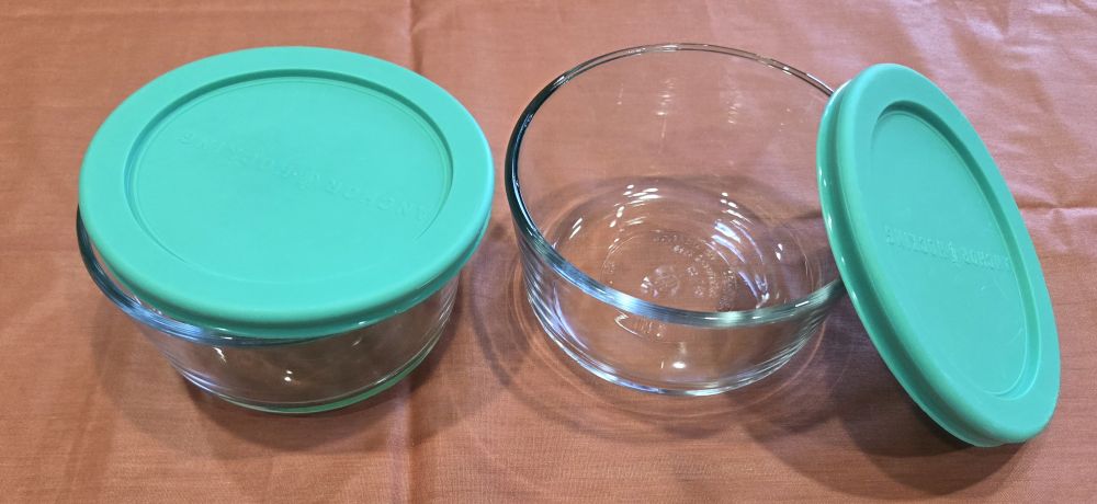 A couple of clear glass food storage bowls with aqua-coloured lids rest atop a coral-coloured cloth surface. Household products such as these are great to have in the kitchen for safely storing hot foods.
