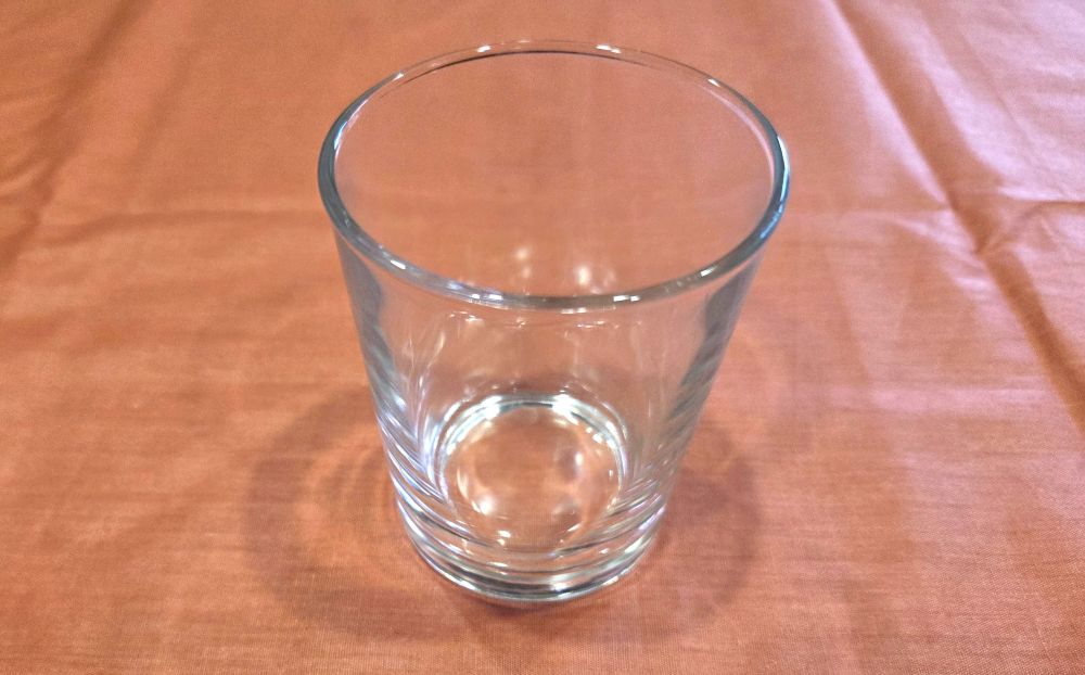 Close-up view of a clear glass five-ounce toddler-sized glass resting atop a coral-coloured fabric on the counter. Glass household products for the kitchen are the safest way to drink beverages toxin-free.