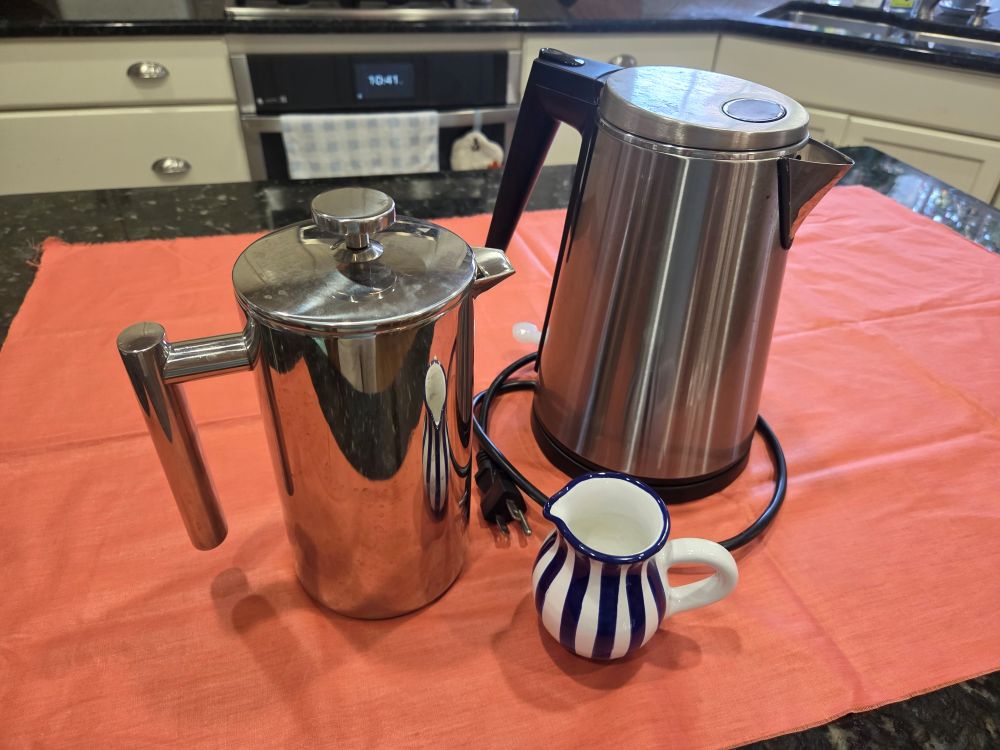A 34-ounce stainless steel coffee press, small blue and white striped ceramic cream pitcher, and stainless steel electric water kettle sit atop a coral-coloured fabric swath on a kitchen counter