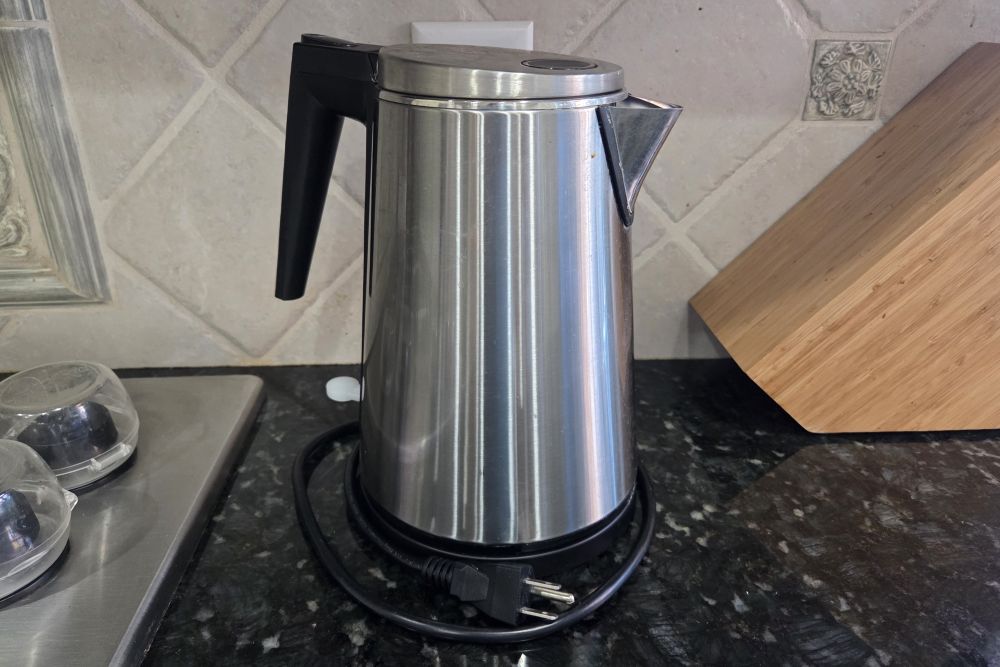 A stainless steel electric water kettle is on a kitchen counter between the edge of the stovetop and knife block. Household products used to prepare food or beverages such as this should never be made out of plastic since it boils water