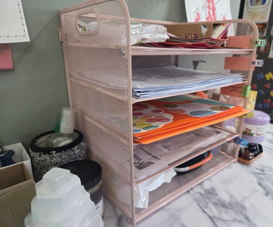 Pink five-tier metal mesh paperwork organizer tray filled with papers and various items, viewed diagonally from the front/side, atop a table alongside various other items