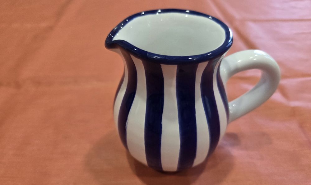 A very cute dark blue and white-striped painted ceramic cream pitcher with tiny pouring spout sits atop a coral-coloured fabric surface