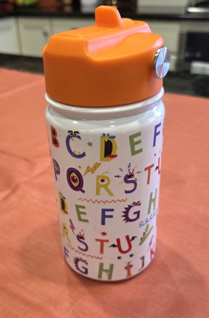 A children's stainless steel water bottle with colourful 'alphabet monsters' and a straw lid (flip-lid tucked in closed position) rests atop a coral-coloured fabric on the counter