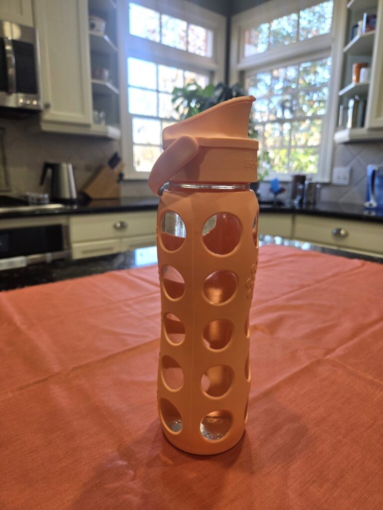 A clear 22-ounce glass water bottle with a flip cap spout for drinking and a peach/orange-coloured silicone sleeve with a circle hole pattern sits atop a coral-coloured fabric swatch on a kitchen counter. Having toxin-free household products like a glass water bottle is a great way to ensure plastics aren't leaching into your water over the course of the day/days.