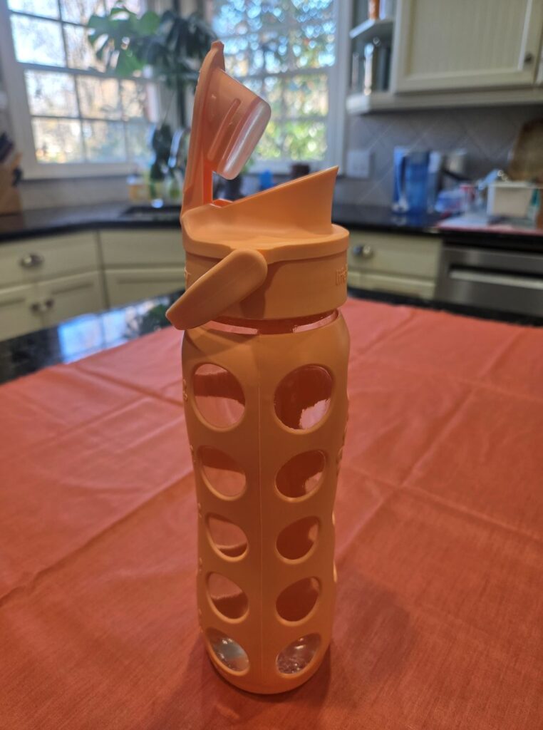 Seen is a clear 22-ounce glass water bottle with its flip cap spout propped open. Its peach/orange-coloured silicone sleeve with a circle hole pattern protects it from accidental drops. Toxin-free household products like this water bottle support a healthy lifestyle since you can drink water all day and not worry about plastics leaching into it.
