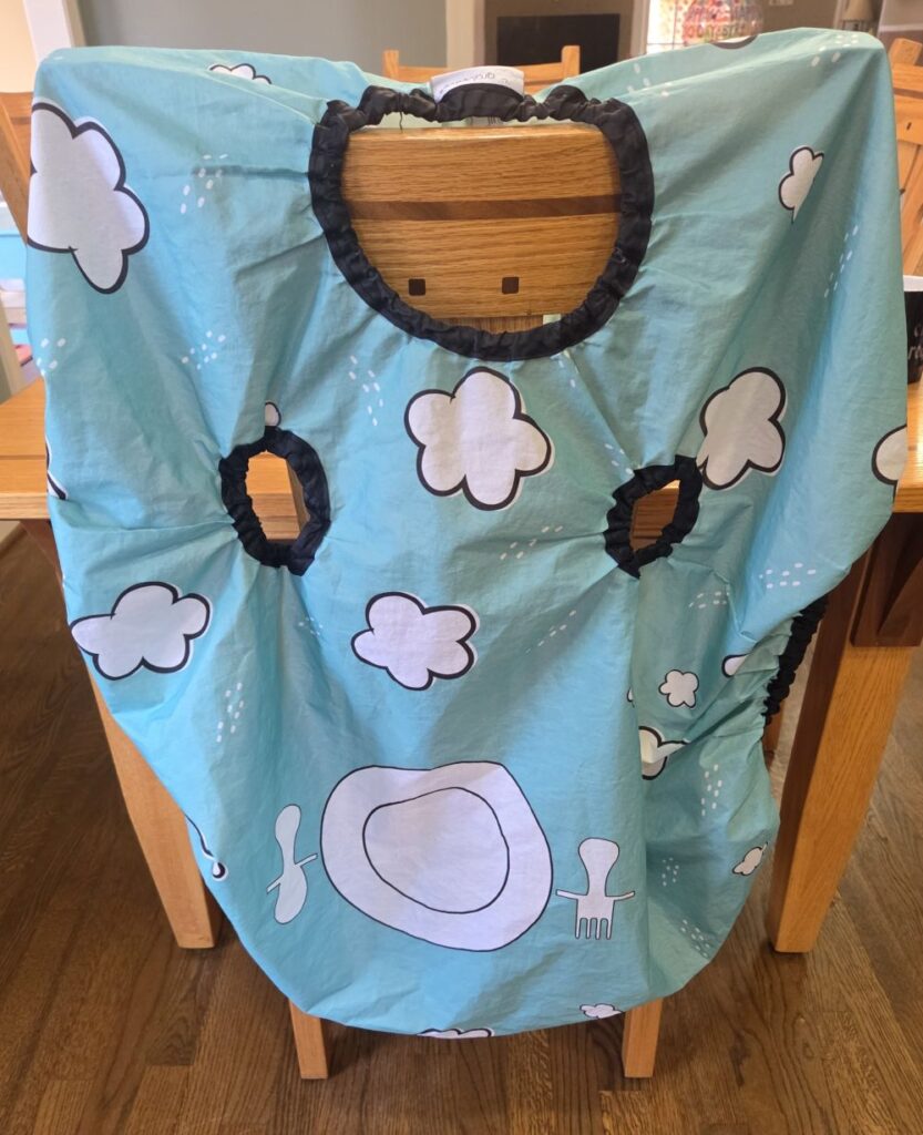 A large waterproof bib smock, with a neck hole and two arm holes, hangs on the back of a kitchen chair. It's blue with a picture of a plate and utensils in white along with white clouds