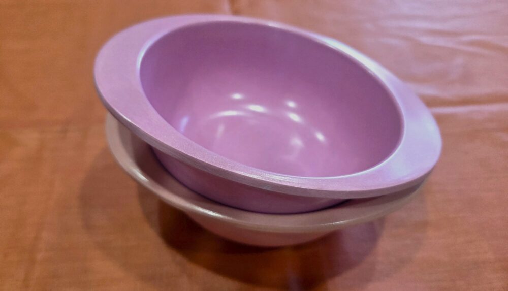 Two nested toddler bowls (in purple and orange) made out of bamboo sit atop a counter draped in a coral cloth