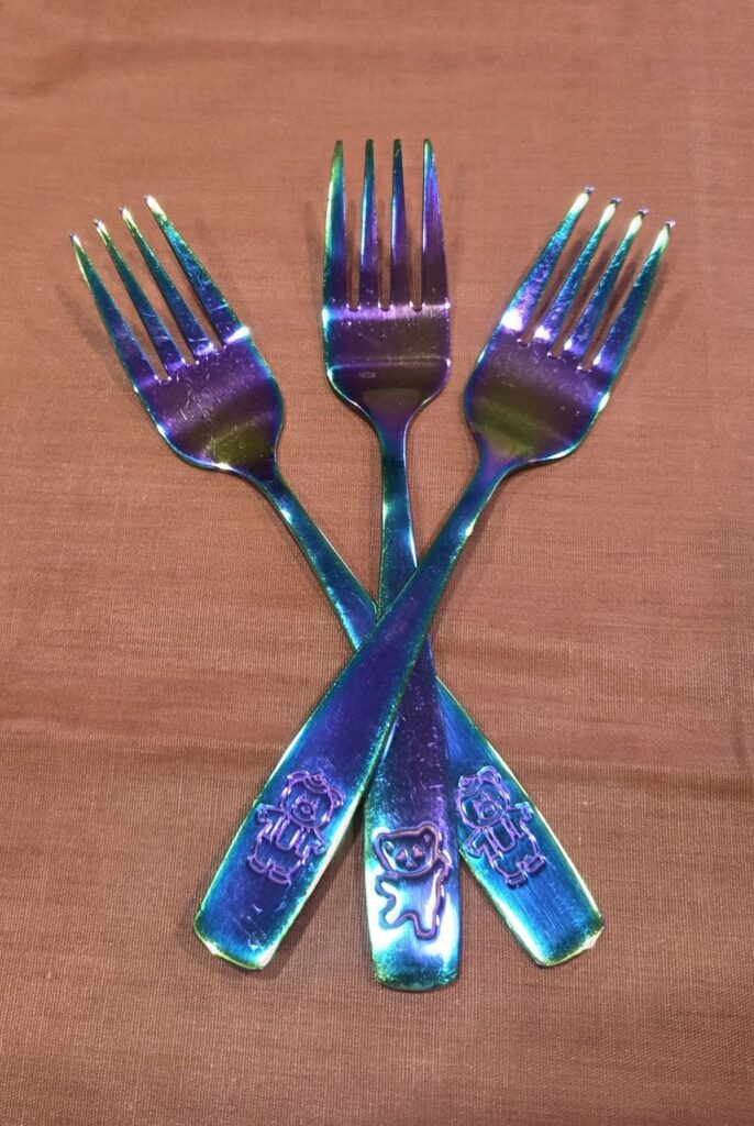 Three iridescent-coloured stainless steel toddler forks are arranged crossed overtop of each other atop a coral-coloured cloth on the counter