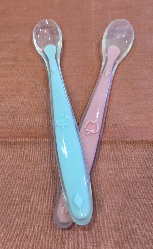 Two silicone toddler spoons, in pale blue and pale pink, are arranged crossed overtop of each other atop a coral-coloured cloth on the counter