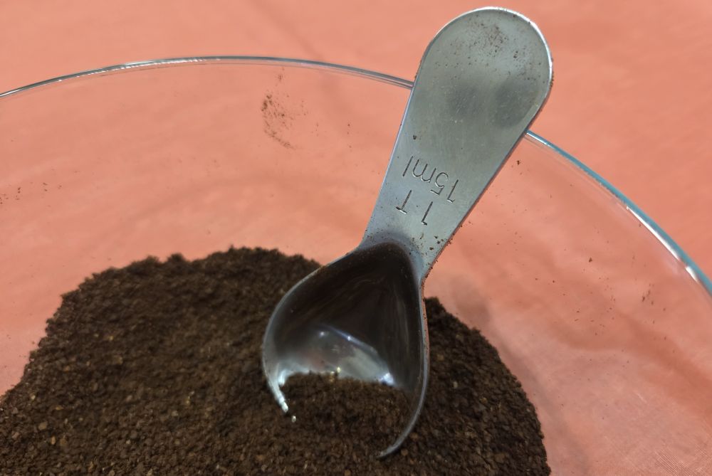 Close-up photo of a stainless steel tablespoon resting inside a clear glass bowl filled with coffee grinds