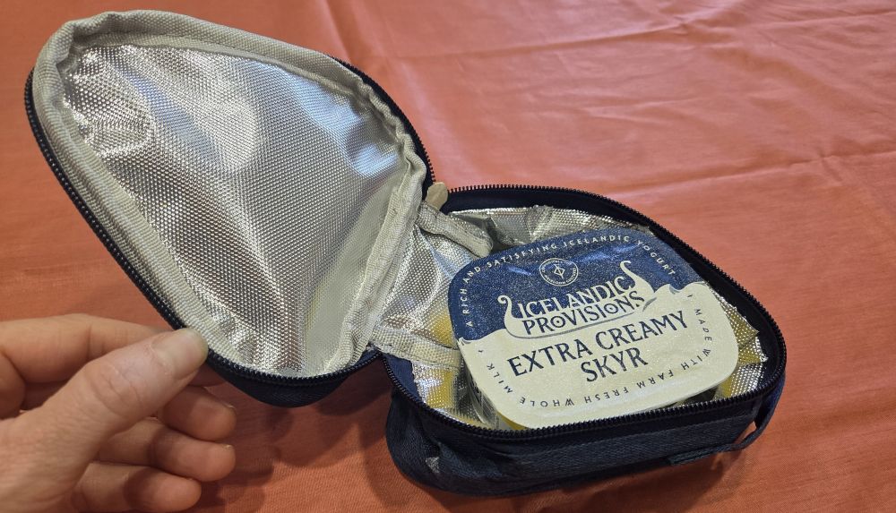 Close-up photo of a soft blue triangle-shaped insulated food storage container with a lid unzipped and being held open to reveal its reflective silver insulated lining inside and a packet of delicious yoghurt