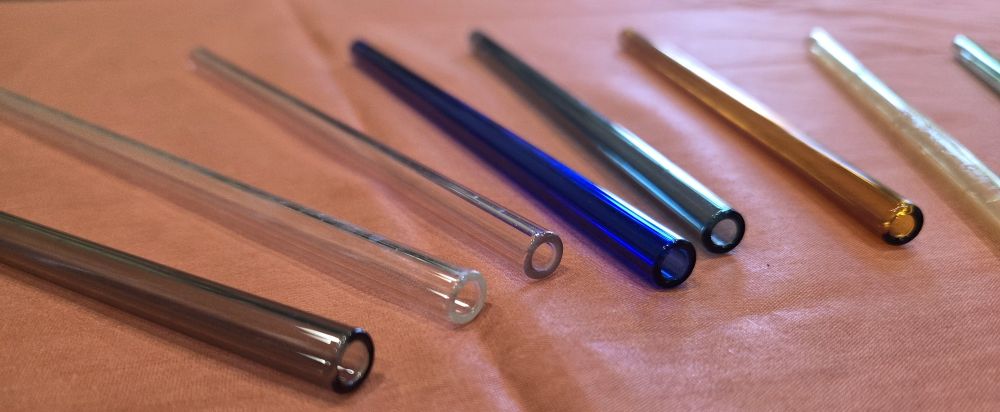 Several different coloured clear glass straws are laid flat in a row atop a coral-coloured fabric surface