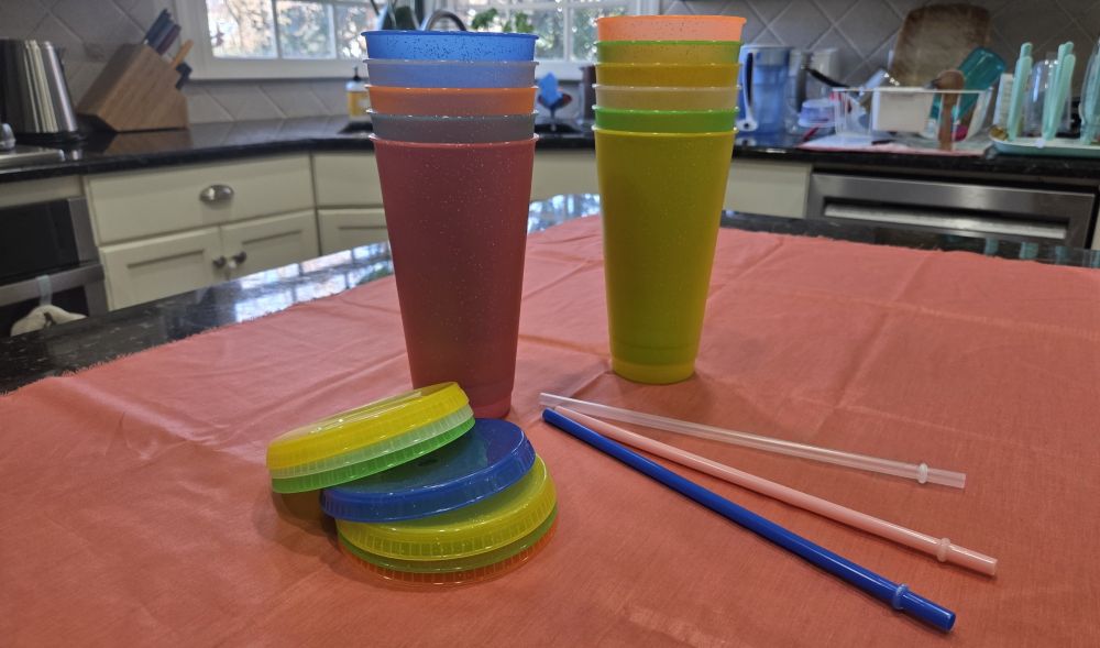 Two stacks of sparkly plastic tumblers in bright colours, along with matching lids and straws, sit atop a coral-coloured cloth on a kitchen counter. These are great household products to have when you want to have a spill-free party because the lids and straws keep the bevvies contained.