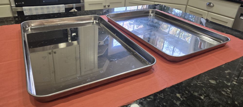 Photo of two large stainless steel baking sheets atop a coral-coloured fabric swath on a countertop. Kitchen household products such as these make baking toxin-free since stainless steel is one of the safest cooking materials.